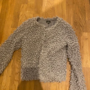 Grå teddyjacka från KappAhl - Mysig grå teddyjacka från KappAhl med långärmad design. Perfekt för kyliga dagar med sitt fluffiga material som ger en trendig look. Jackan har en enkel och stilren stil utan krage.