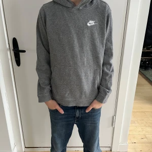 Grå hoodie från Nike - Säljer en grå huvtröja från Nike i storlek L (147–158 cm). Tröjan har en klassisk design med en liten Nike-logga på bröstet och en känguruficka framtill. Perfekt för vardag eller träning.  Personen på bilderna är 12 år gammal, 160 cm lång och har en smal/normal kroppsbyggnad.  Skick: Bra begagnat skick med några tecken på användning.  Nypris: 600 SEK