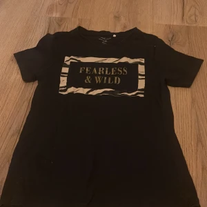 Svart t-shirt från Name It - Cool svart t-shirt från Name It med texten 'Fearless & Wild' i ett zebramönstrat tryck på framsidan. Perfekt för den modiga och vilda! Kortärmad och bekväm passform.