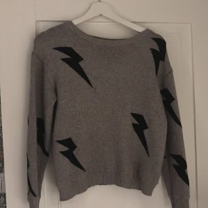 Grå/beige stickad tröja  - Snygg grå/begie  tröja med svarta blixtar. Tröjan har en rund hals och långa ärmar, perfekt för en cool och avslappnad stil. Den är gjord i ett mjukt material som känns skönt mot huden.