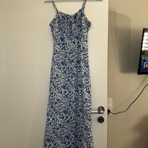 Blåblommig långklänning med smala axelband - Charmig långklänning med blått blommönster på vit bakgrund. Klänningen har smala axelband och en söt knytning framtill. Perfekt för somriga dagar.