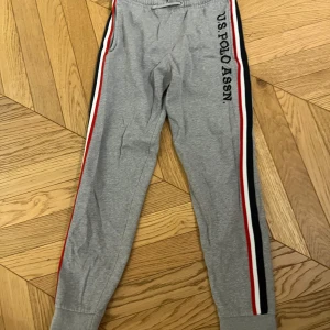 Grå mjukisbyxor från U.S. Polo Assn. - Snygga grå mjukisbyxor från U.S. Polo Assn. med elastisk midja och snörning. Byxorna har en cool randig detalj i rött, vitt och svart längs sidorna samt en ficka. Perfekta för en avslappnad stil.
