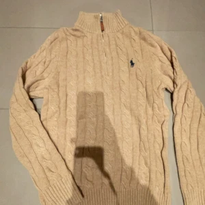 Beige stickad tröja från Polo Ralph Lauren - Säljer en stilren beige stickad tröja från Polo Ralph Lauren med kabelstickat mönster och en liten broderad logga på bröstet. Tröjan har en dragkedja vid halsen och långa ärmar. Perfekt för kyliga dagar!