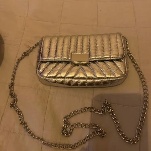 Silverfärgad quiltad axelväska - Snygg silverfärgad axelväska med quiltad design och kedjerem. Perfekt för att ge din outfit en glittrig touch. Väskan har en stilren rektangulär form och en praktisk storlek för att bära dina nödvändigheter.