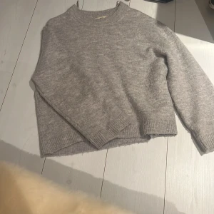 Grå stickad tröja från Gina Tricot - Mysig grå stickad tröja från Gina Tricot i premiumkvalitet. Perfekt för kyliga dagar med sin klassiska design och ribbade detaljer vid hals, ärmslut och nederkant.