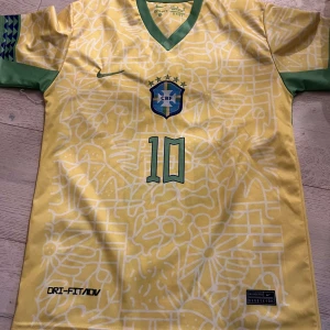 Gul Brasilien fotbollströja från Nike - Säljer en gul Brasilien fotbollströja från Nike med gröna detaljer och nummer 10 på framsidan. Tröjan har korta ärmar och är tillverkad i Dri-FIT material för optimal komfort. Perfekt för fotbollsfans!