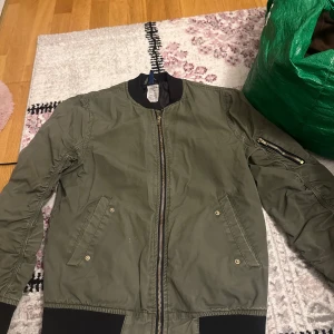 Olivgrön bomberjacka från H&M - Säljer en olivgrön bomberjacka från H&M med dragkedja och ribbade muddar. Jackan har en klassisk bomberstil med fickor framtill och en ärmficka med dragkedja. Perfekt för en cool och avslappnad look.