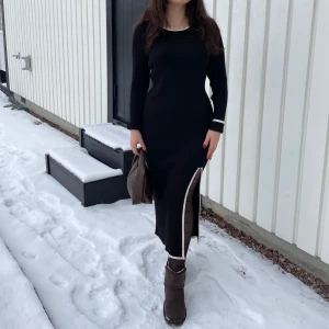 Svart långklänning med slits M nyskick  - Elegant svart långklänning med vita detaljer och en stilren slits. Klänningen har långa ärmar och en figurnära passform, perfekt för kyligare dagar.