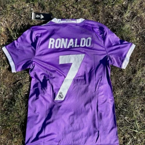 Lila Real Madrid fotbollströja - Säljer en lila Real Madrid fotbollströja från Adidas med 'Ronaldo' och nummer 7 på ryggen. Tröjan har korta ärmar och vit krage med vita detaljer. Perfekt för fans av Ronaldo och Real Madrid! 💜
