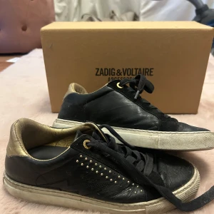 Svarta sneakers från Zadig & Voltaire - Snygga svarta sneakers i skinn från Zadig & Voltaire med gulddetaljer och snörning. Skorna har en cool blixtdetalj på sidan och små nitar för extra edge. I använt skick men har absolut frtf mer att ge! Går garanterat att snygga till dom och fräscha upp dom 🌟❤️ Passar på mig som brukar ha 38 - 39 