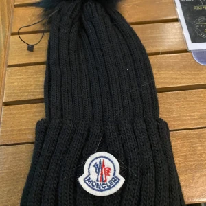 Svart stickad mössa från Moncler - Snygg svart stickad mössa från Moncler med en fluffig tofs på toppen. Mössan har en broderad logotyp framtill och är perfekt för kyliga dagar.