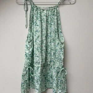 Grön blommig ärmlös topp - Säljer en söt grön blommig topp med knytband vid axlarna. Toppen är ärmlös och har en luftig passform med volangdetaljer vid midjan. Perfekt för varma sommardagar!