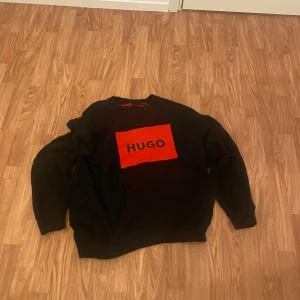 Svart tröja från Hugo Boss - Snygg svart tröja från Hugo Boss med ett stort rött tryck på bröstet. Tröjan har långa ärmar och en klassisk passform. Perfekt för en stilren look.