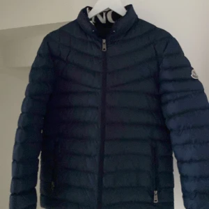 Mörkblå dunjacka från Moncler - Snygg mörkblå dunjacka från Moncler med dragkedja och broderad logga på ärmen. Perfekt för kyliga dagar med sin quiltade design som håller värmen. Jackan har en stilren look och passar bra till olika outfits. Priset kan diskuteras.
