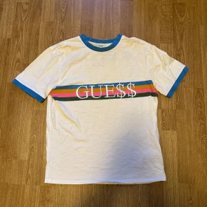 Vit t-shirt Guess x A$AP Rocky - Snygg vit t-shirt från Guess med färgglada ränder och logga på framsidan. Blå detaljer vid krage och ärmslut ger en fräsch look. Lite oversized. Röd tråd för att sy ihop kragen. Ej gjort av mig men sänker priset pga detta.