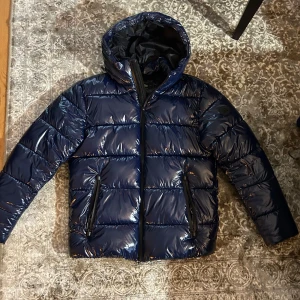Blå pufferjacka från Carrera - Säljer en mörkblå glansig pufferjacka från Carrera i storlek medium. Jackan har en glansig yta och är utrustad med en huva och dragkedja framtill. Perfekt för kyligare dagar. Nypris på jackan ligger på 1900kr och mitt pris är 799kr. Helt ny jacka och knappt använd! Hör av di vid fler funderingar eller fler bilder! 🙌