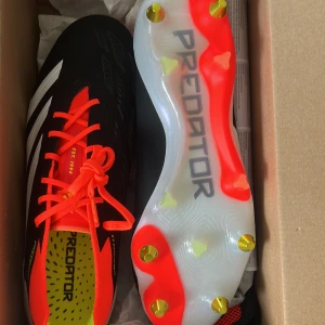 Adidas Predator Elite SG Fotbollsskor - Säljer ett par Adidas Predator Elite SG fotbollsskor i svart och röd med vita detaljer. Skorna har en robust design med snörning och är perfekta för spel på mjuka underlag. De har en iögonfallande sula med röda och gula detaljer.