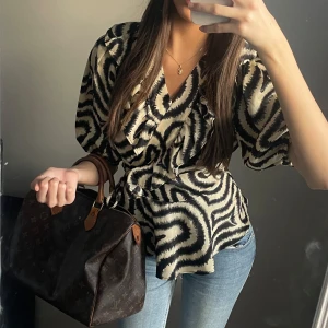 Unik omlottblus - As Snygg omlottblus med svart och beige mönster. Blusen har korta ärmar och volangdetaljer längs halsringningen. Perfekt för en trendig look.