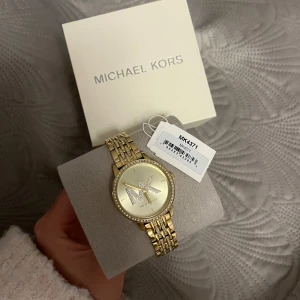 Guldfärgad klocka från Michael Kors - Snygg guldfärgad klocka från Michael Kors med glittriga detaljer runt urtavlan och ett stort MK-emblem.  Använd 1-2 gånger, superbra skick inga repor eller liknande. Boks, lappar och länk för att förlänga den finns kvar. Batteriet måste bytas. Köpt för 3800