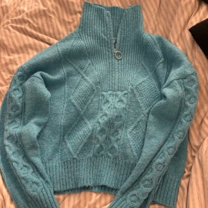 Turkos stickad tröja med dragkedja - Mysig turkos stickad tröja med hög krage och dragkedja. Tröjan har ett fint flätmönster och ribbade muddar vid ärmslut och nederkant. Perfekt för kyliga dagar!