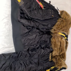 Svart bomberjacka med pälsdetaljer från Alpha Industries - Säljer en svart bomberjacka från Alpha Industries med pälsdetaljer på huvan. Jackan har en gul insida och flera fickor. Perfekt för kyligare väder.