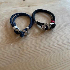 Två armband med ankardetalj - Säljer två snygga armband med ankardetalj. Ett i marinblått med vita detaljer och ett i svart med röda detaljer. Perfekta för en stilren look.