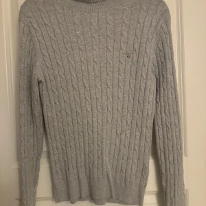 Grå kabelstickad tröja från Gant - Säljer en stilren grå kabelstickad tröja från Gant. Tröjan har en klassisk rund hals och långa ärmar. Perfekt för kyligare dagar och ger en tidlös look.