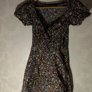 Blommig klänning med snörning - Charmig blommig klänning från Bikbok med korta ärmar och snörning framtill. Klänningen har ett färgglatt mönster med blommor i olika nyanser av rosa, blått, gult och orange på en mörk bakgrund. Perfekt för en somrig look!