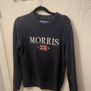 Mörkblå tröja från Morris - Snygg mörkblå tröja från Morris med broderad logotyp och Union Jack-flagga på bröstet. Tröjan har långa ärmar och ribbade muddar vid hals, ärmslut och nederkant. Perfekt för en stilren look. Passar även S! 