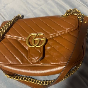 Brun quiltad axelväska med kedja - Snygg brun axelväska i skinn med quiltad design och guldfärgad kedja. Väskan har ett stort GG-spänne framtill och en praktisk innerficka. Perfekt för att ge din outfit en lyxig touch.
