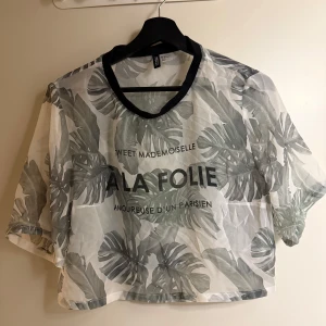 Transparent bladmönstrad topp från H&M - Säljer en transparent topp från H&M Divided med grönt bladmönster och texten 'A LA FOLIE' på framsidan. Toppen har korta ärmar och en svart kant vid halsringningen. Perfekt för en trendig look!