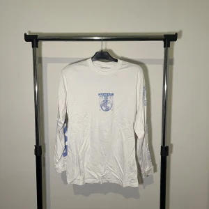 Vit långärmad tröja från Vailent - Säljer en vit långärmad tröja från Vailent med blått tryck fram och bak. Tröjan har ett coolt grafiskt mönster med texten 'Hysteria' och en jordglob. Perfekt för en avslappnad stil.