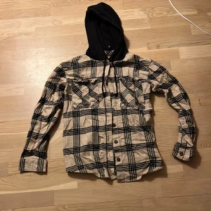 Rutig skjorta med huva från HM  - Snygg rutig skjorta från HM med svart huva. Skjortan har långa ärmar och knappar framtill. Perfekt för en avslappnad stil.