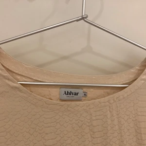 Beige topp från Ahlvar Gallery - Säljer en elegant beige topp från Ahlvar Gallery med ett subtilt mönster. Toppen har trekvartsärmar och knappar vid ärmsluten. Perfekt för en stilren look.