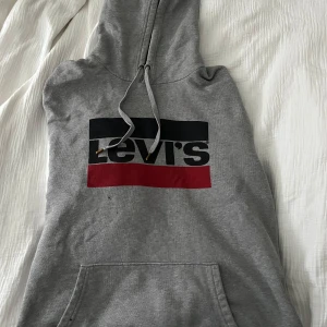 Grå hoodie från Levi's - Säljer en grå hoodie från Levi's med en stor logga i svart och rött på framsidan. Tröjan har en klassisk känguruficka och justerbar huva med dragsko. Perfekt för en avslappnad stil.