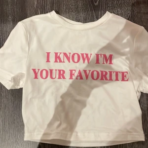 Vit t-shirt med rosa text - Säljer en vit t-shirt med korta ärmar och trycket 'I KNOW I'M YOUR FAVORITE' i rosa på framsidan. Perfekt för en avslappnad stil. Iconic!!!
