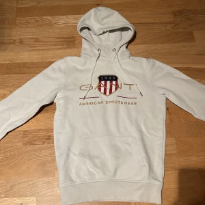 Vit hoodie från GANT - Säljer en stilren vit hoodie från GANT med broderad logga och texten 'American Sportswear' på framsidan. Hoodien har en praktisk ficka framtill och justerbar huva med snören.