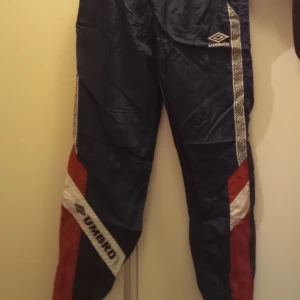 Blå träningsbyxor från Umbro - Säljer ett par klassiska blå träningsbyxor från Umbro med elastisk midja och vita samt röda detaljer längs sidorna. Byxorna har en sportig look med Umbro-loggan på benet och mönstrade band längs sidorna.
