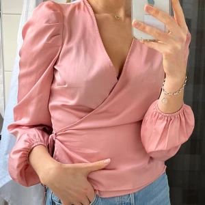 Rosa omlottblus från Bik Bok - Säljer en söt rosa omlottblus från Bik Bok. Blusen har långa ärmar med puffiga axlar och elastiska ärmslut. Den är v-ringad och har en justerbar knytning i sidan för en smickrande passform.