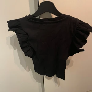 Svart topp med volangärmar från Zara - Säljer en svart topp från Zara med korta volangärmar. Perfekt för en stilren look. Tillverkad i Bangladesh.