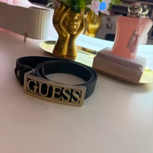 Svart bälte från Guess - Snyggt svart bälte från Guess med ett stort, iögonfallande spänne i guld med märkesnamnet. Perfekt för att ge en stilren touch till din outfit. Bältet är tillverkat i ett elegant material som ger en lyxig känsla.