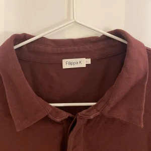 Brunröd pikétröja från Filippa K - Snygg brun pikétröja från Filippa K med klassisk krage och korta ärmar. Perfekt för en stilren och avslappnad look. Tveka inte att hojta till vid eventuella frågor!