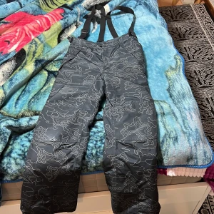 Svarta vinterbyxor med hängslen - Säljer ett par svarta vinterbyxor med justerbara hängslen och ett coolt abstrakt mönster. Perfekta för kalla dagar med sin varma och fodrade insida. Byxorna har en praktisk design med knäppning vid bensluten.