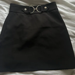 Svart kjol från Amisu - Snygg svart kjol från Amisu med dekorativa ringdetaljer framtill och en dragkedja bak. Aldrig använd men lappen är ej kvar.🫶🏻Hör av er vid frågor. ☺️
