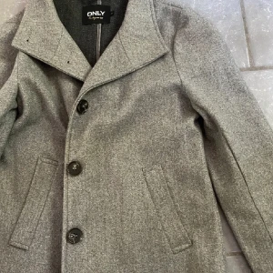 Grå kappa från ONLY - Snygg grå kappa från ONLY med klassisk design och stora knappar framtill. Köpt för 399kr säljer för 199kr. Endast använd fåtal gånger, ser nästan ut som ny! 