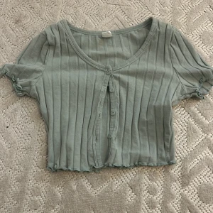 Grön ribbad kortärmad topp - Säljer en söt grön ribbad topp med korta ärmar och knappar framtill. Toppen är croppad och har en u-ringad design med små volangdetaljer vid ärmslut och nederkant.