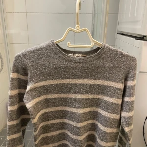 randig tröja från H&M - Säljer en mysig grå tröja med beige ränder från H&M. Tröjan har långa ärmar och en rund halsringning, perfekt för kyligare dagar.