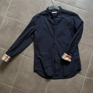 Burberry skjorta - Snygg mörkblå skjorta från Burberry Brit med klassiska rutor på insidan av manschetterna. Skjortan har knappar framtill och en stilren design som passar perfekt för olika tillfällen.
