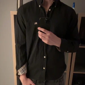 Svart skjorta från Burberry - Snygg svart skjorta från Burberry med klassiskt rutigt mönster på insidan av kragen. Skjortan är köpt på luxury vintage i Rom så den bara vara äkta. Skjortan har tyvärr tappat en knapp då den fastnade i ett bord på en fest. Har även ett litet hål i armhålan