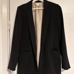 Svart kavaj från H&M - Elegant svart kavaj från H&M med klassisk design. Den har långa ärmar och en stilren passform, perfekt för en chic look. Kavajen är fodrad med ett ljust tyg som ger en fin kontrast.